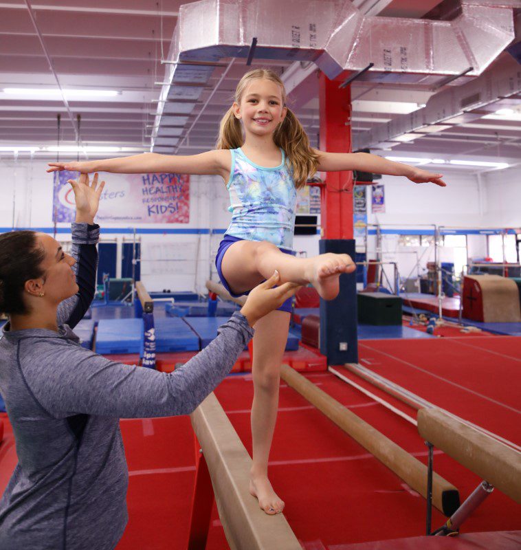 gymnastics classes in Boca Raton, young girl on beam kicking forward with help of coach