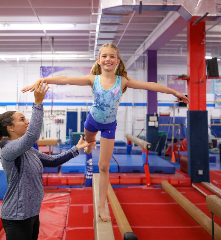 gymnastics classes in Boca Raton, young girl on beam kicking backwards with help of coach gymnastics classes in Boca Raton, young girl on beam kicking backwards with help of coach